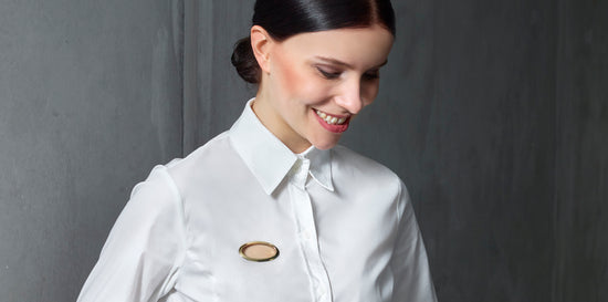Foto einer Frau in einer weißen Bluse. Auf der linken Seite auf Brusthöhe befindet sich ein ovaler vergoldeter Silberrahmen der mit der Bluse vernäht ist. Hemd und Brosche verschmelzen zu einer Einheit und die Trägerin wird zum eigentlichen Schmuckstück.